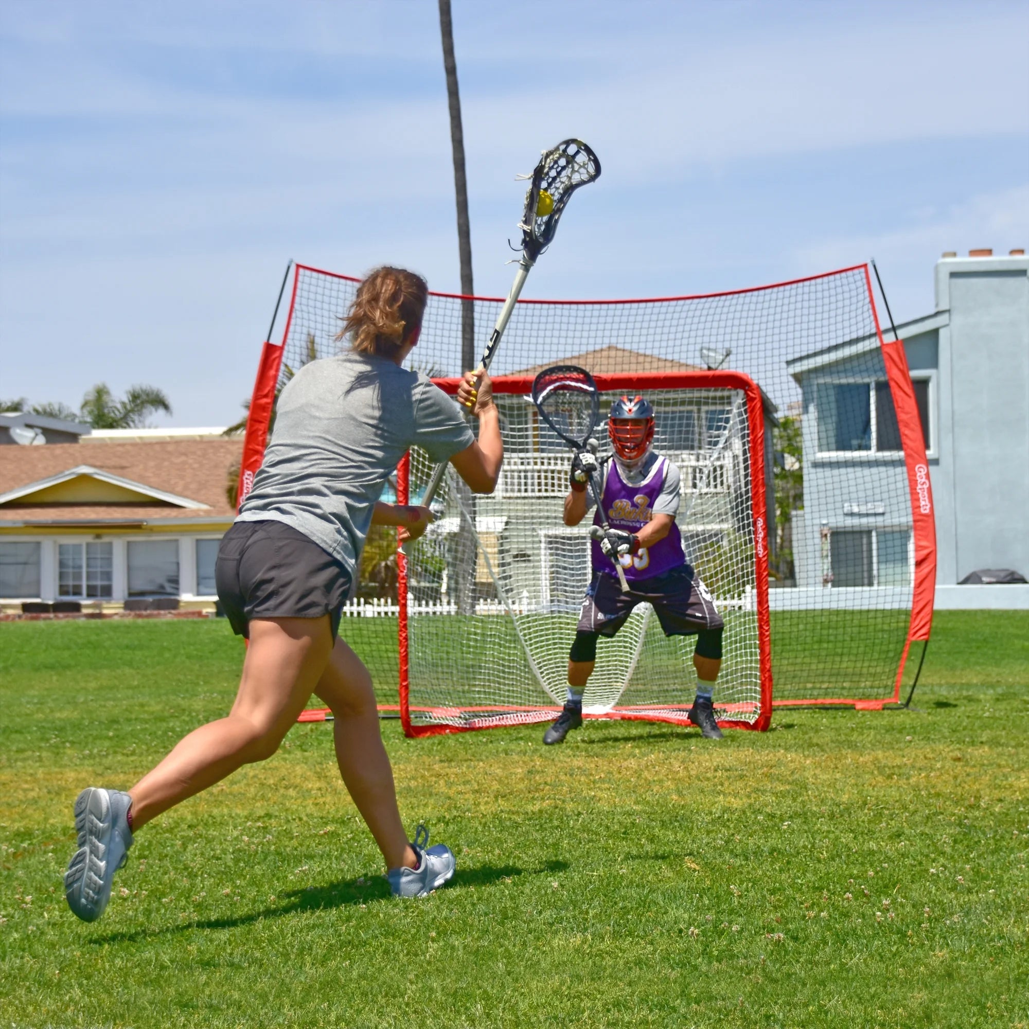 Two people playing lacrosse on a grassy field with a goal in the background.
