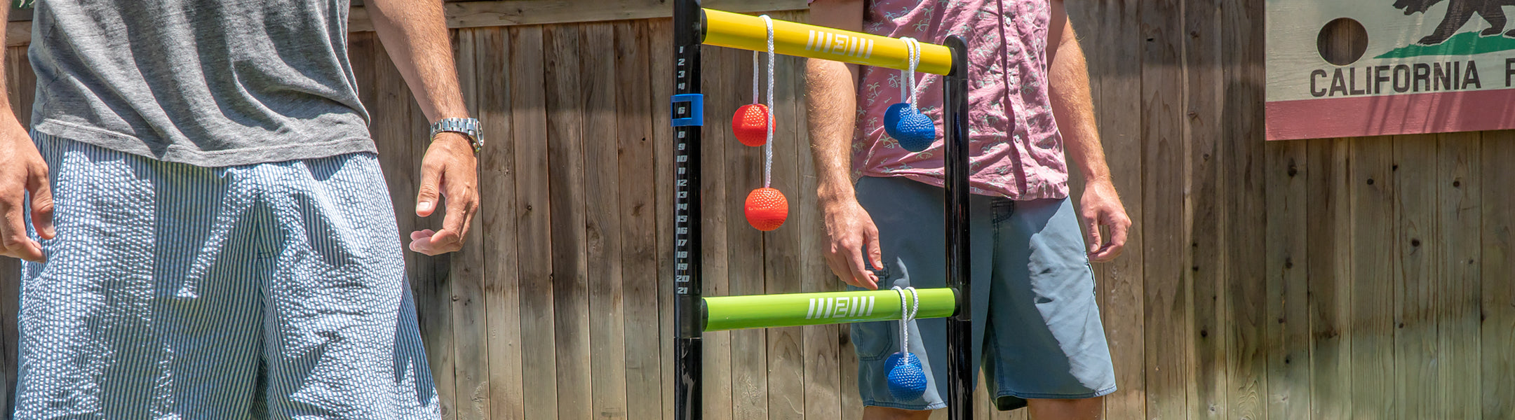 Two people playing with a colorful ladder game outdoors, with a wooden fence and sign in the background.