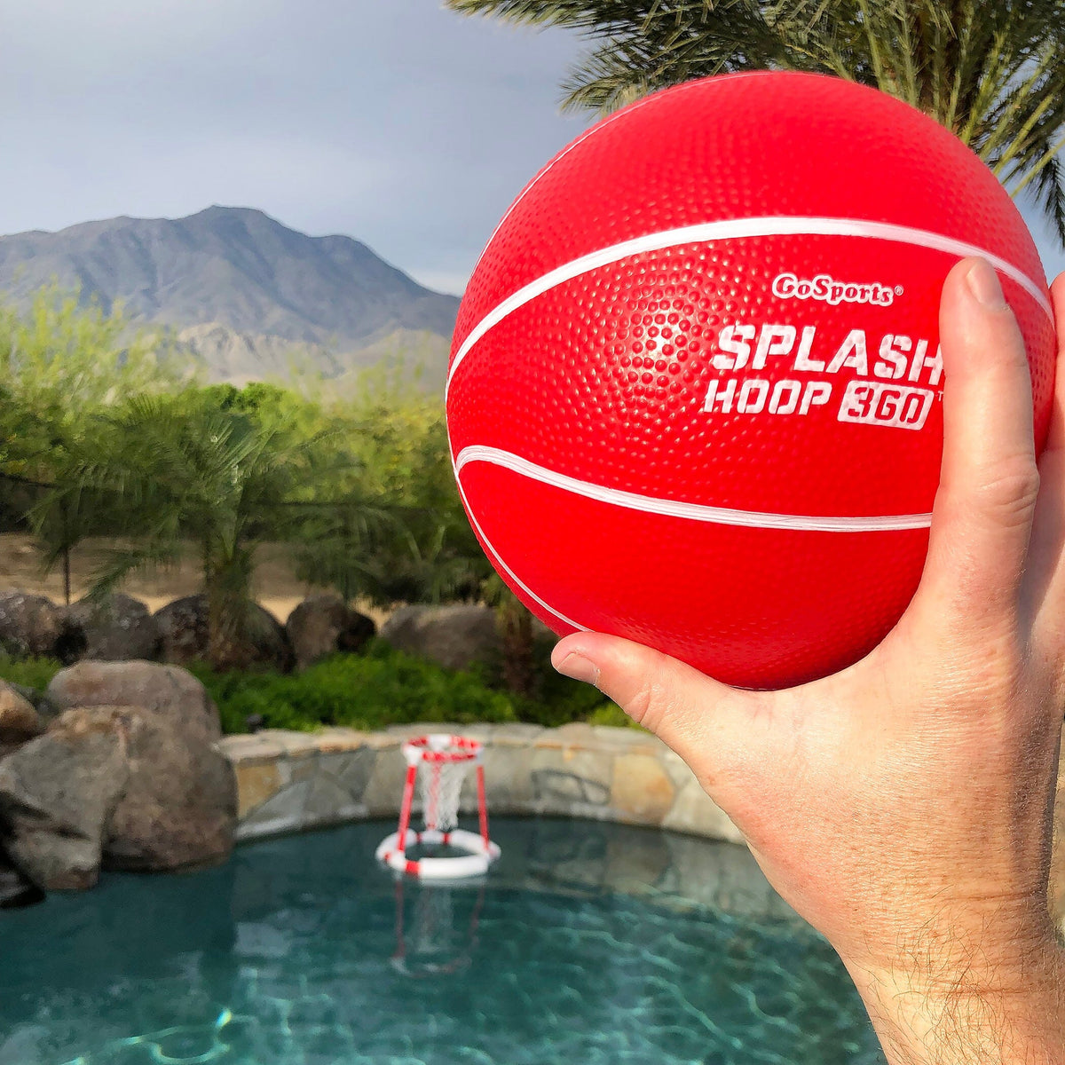 Hand holding a red GoSports Splash Hoop 360 ball, the pool basketball hoop floating in a backyard pool behind