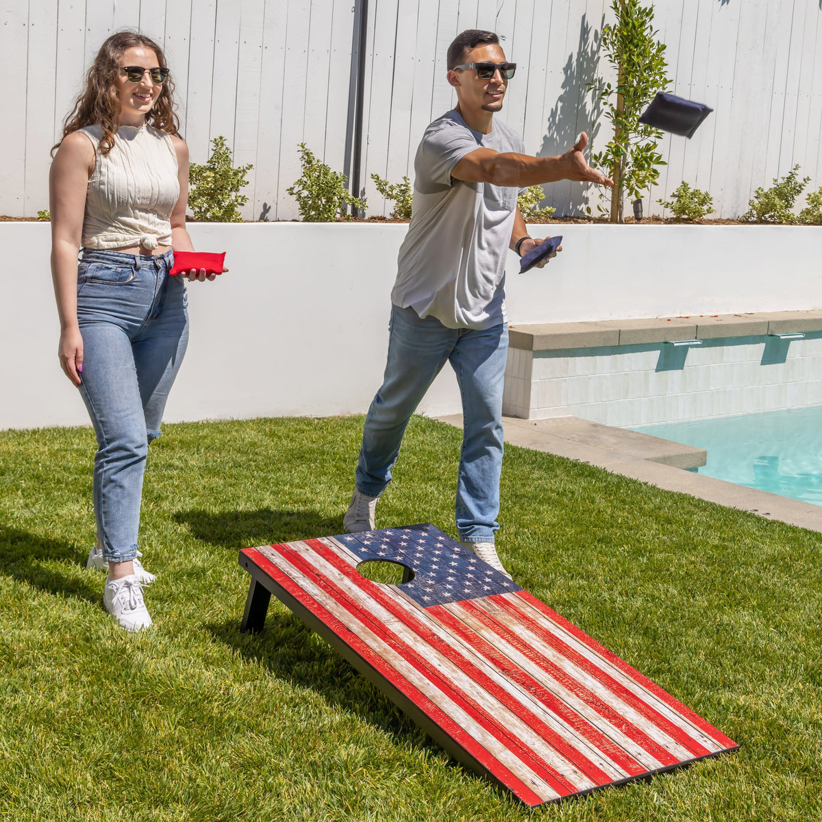 Player tossing a navy bag toward a GoSports American flag cornhole board on a backyard lawn by a pool