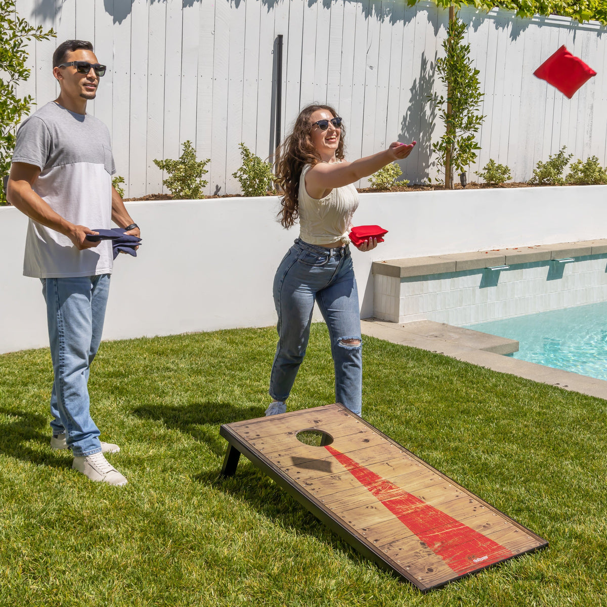 Player tossing a red bag at a rustic wood GoSports cornhole board with red stripe design by a backyard pool