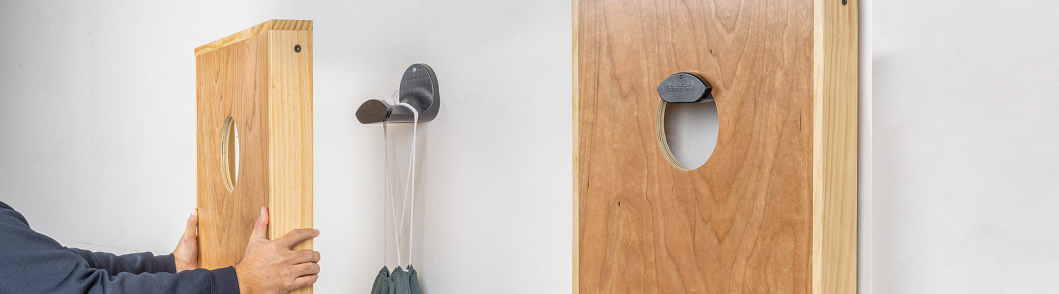 Cornhole board held by a person with hooks on a white wall next to cornhole board hanging from a black hook.