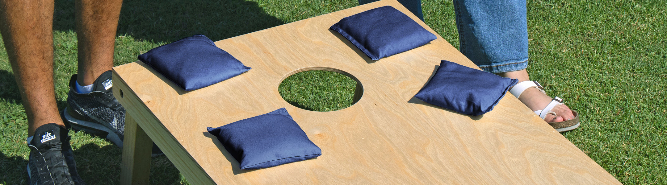 Cornhole game with blue bags on a wooden board, people standing nearby on grass.
