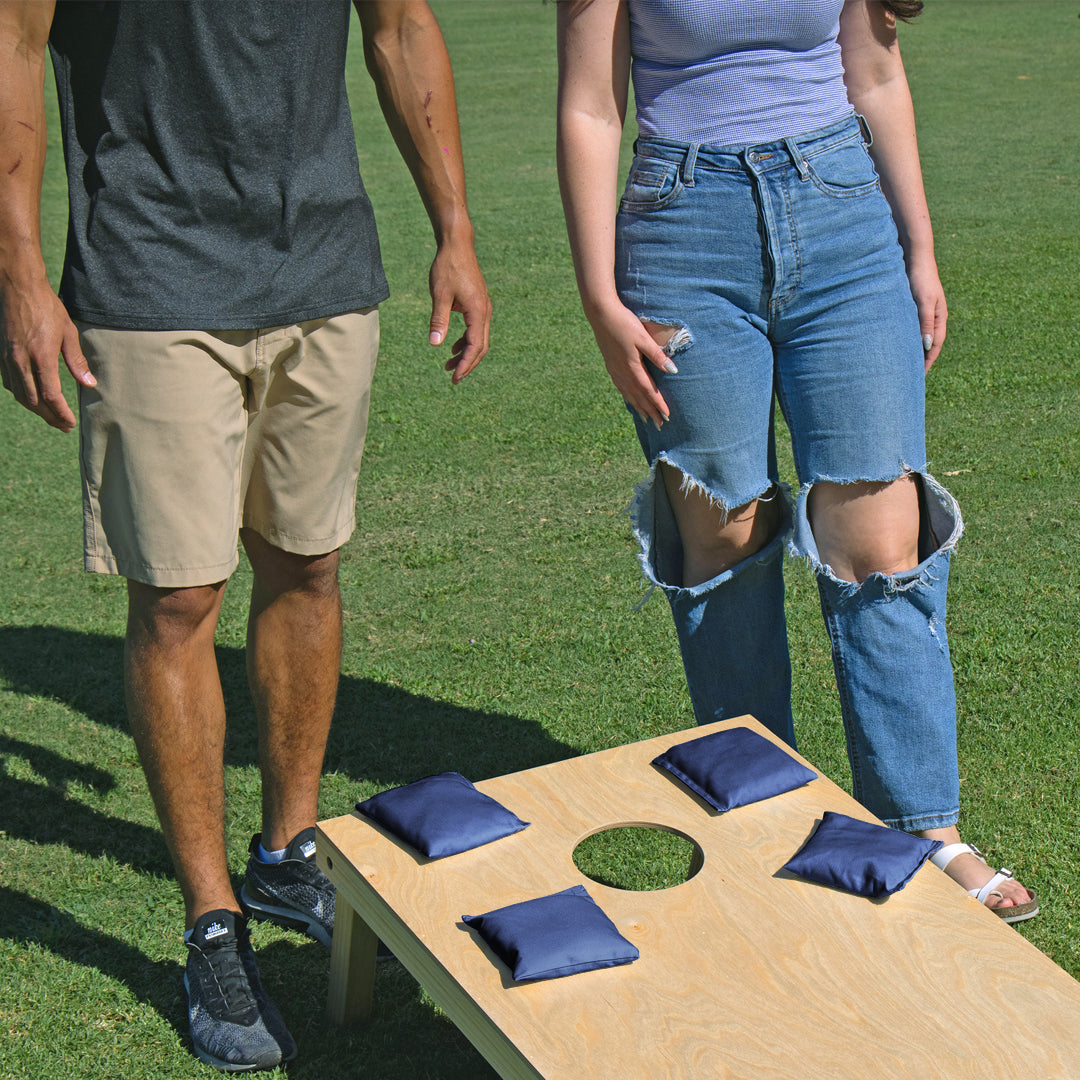 Two people standing on grass next to a cornhole board with blue bags.