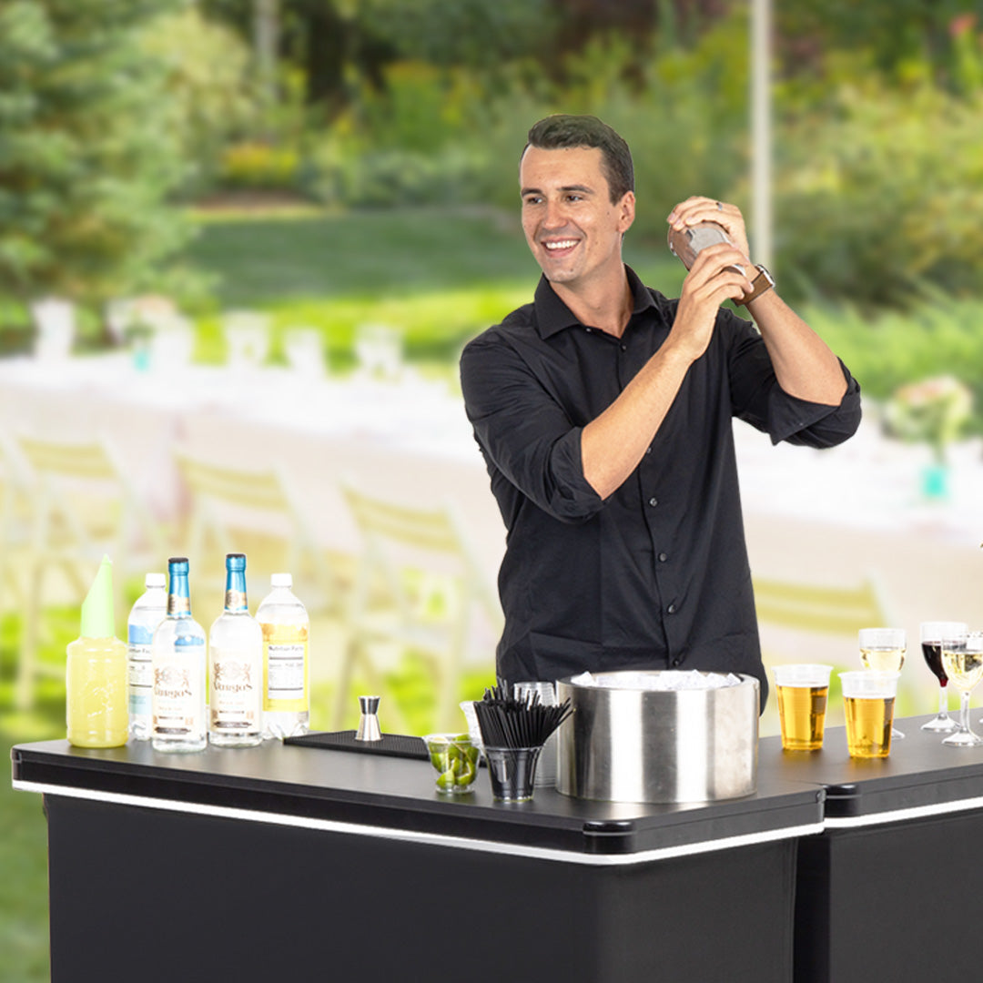 Man making a cocktail at an outdoor bar with bottles and glasses on the counter.