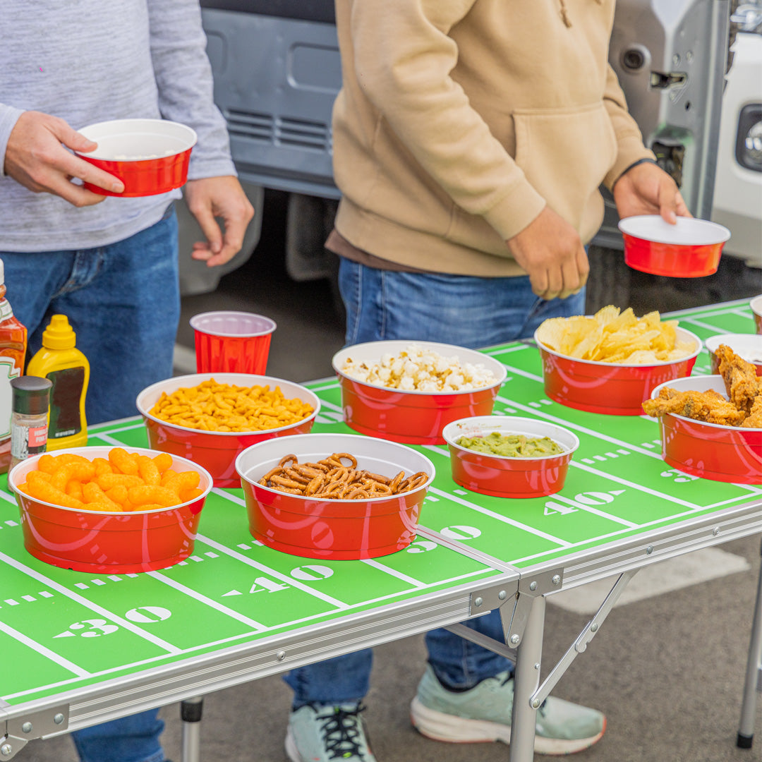 People holding red bowls near a table with snacks on a football field-themed mat.