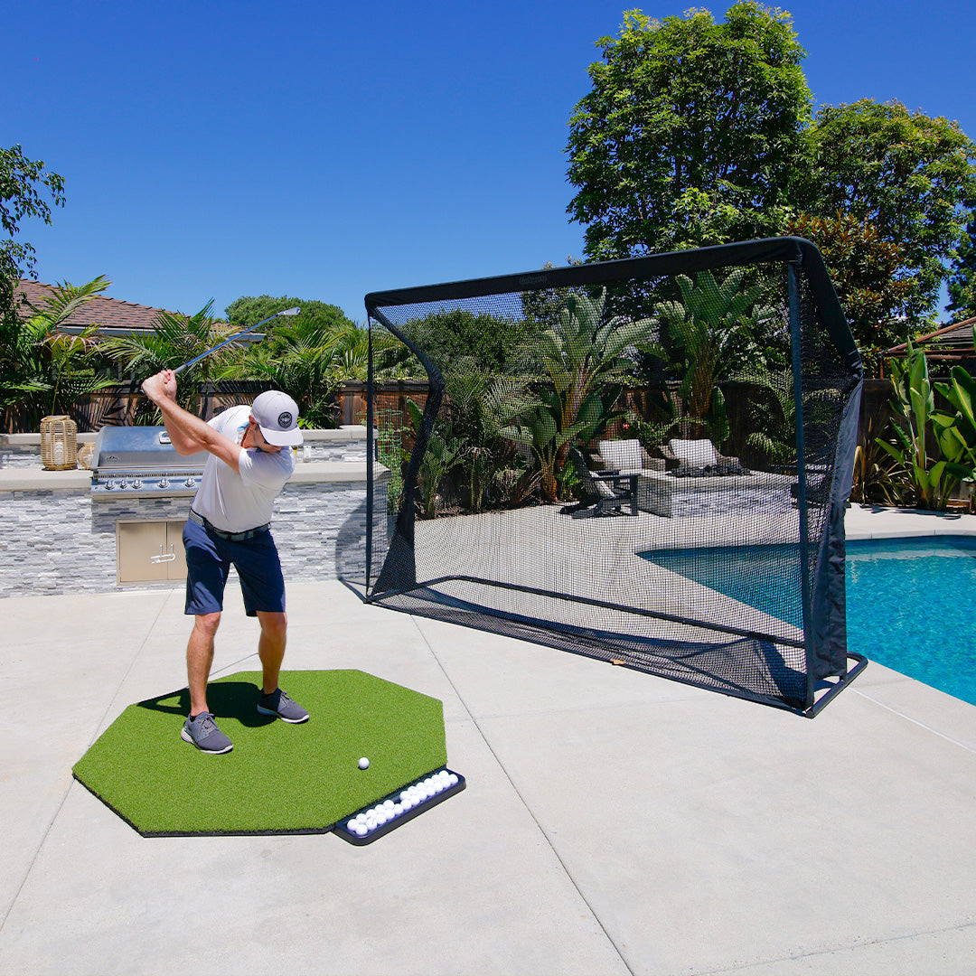 Person practicing golf swings on a mat with a golf practice net in a backyard setting.