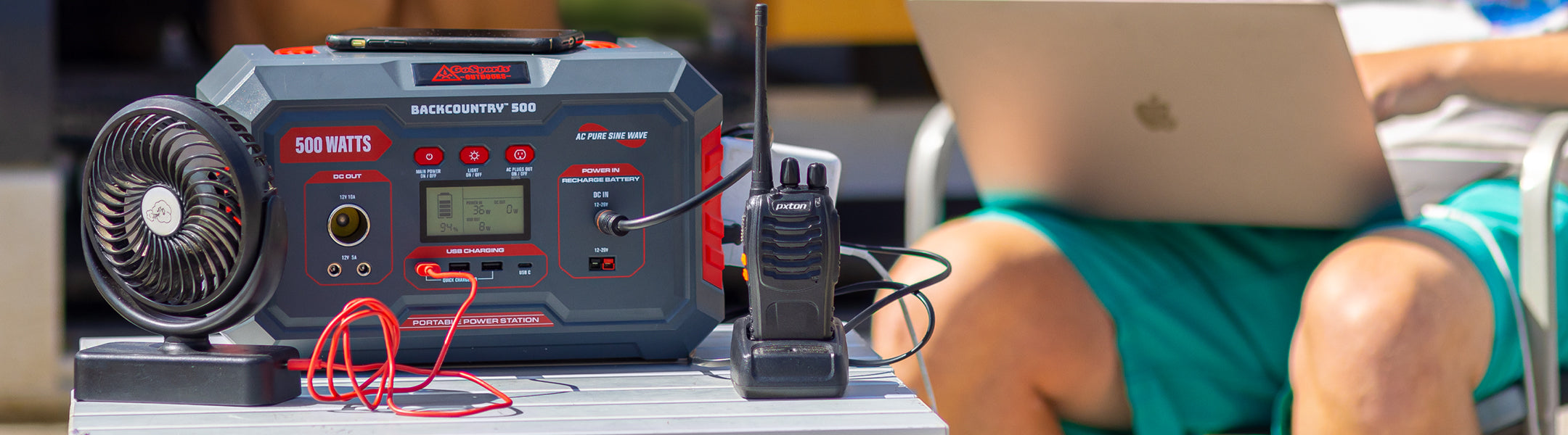 Red and black power station with fan and cables on a table, person using laptop in background