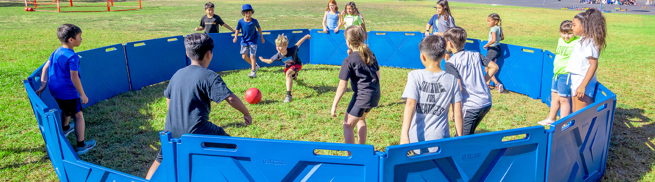 Children playing inside a blue plastic circular panel with a red ball on a grassy field.
