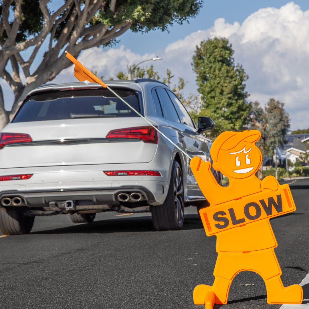 Orange 'SLOW' sign held by a cartoon character in front of a silver car on a road.