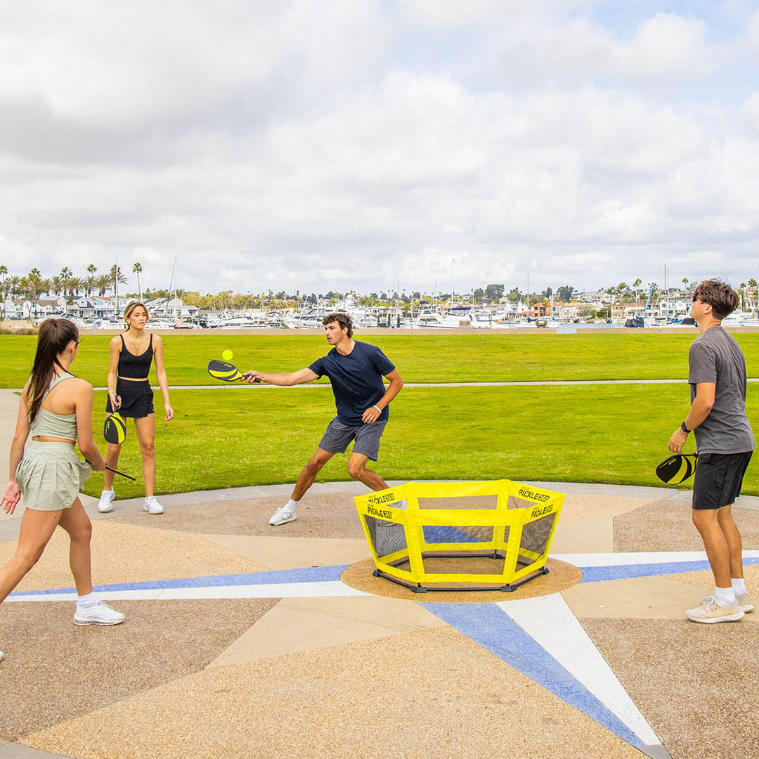 Four people playing with a yellow sports ball and pickleball paddles on a grassy field.