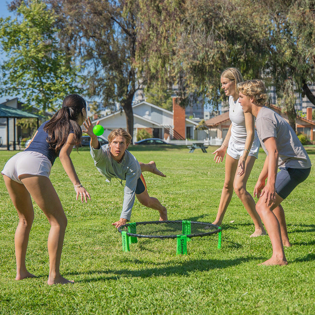 Four people playing slammo roundnet game on a grassy field with a house and trees in the background