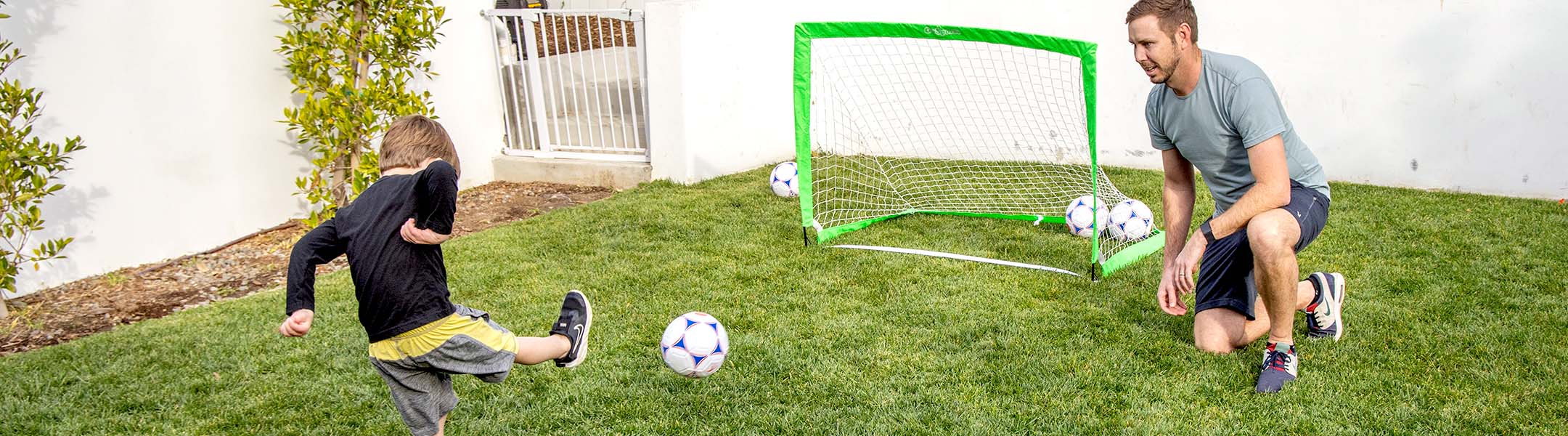 child kicking soccer ball to a green soccer goal with a man standing on a grassy ground