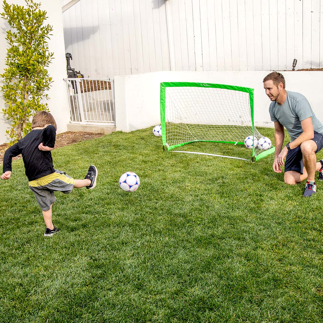 Child kicking a soccer ball towards a green goal with an adult crouching nearby on a grassy backyard.