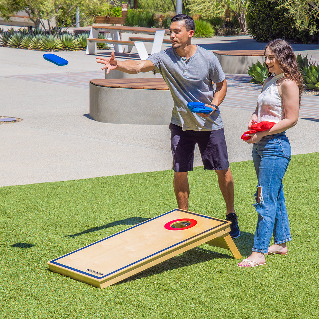 Two people playing a game of cornhole on a grassy area with benches and plants in the background.