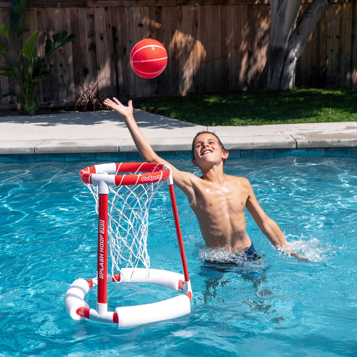 Kid in a pool reaching for a red ball above a floating red & white GoSports Splash Hoop 360 pool basketball hoop