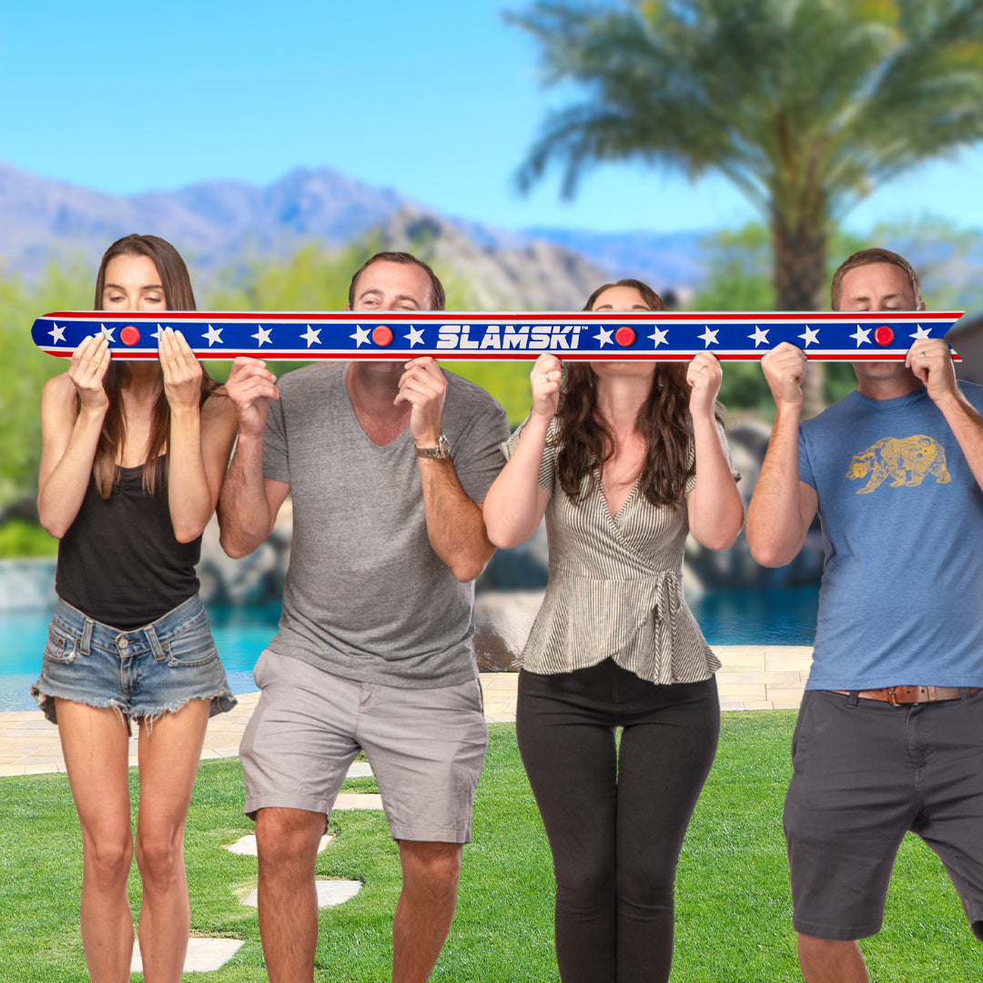 Four people holding a large ski with 'SLAMSKI' branding in a scenic outdoor setting.