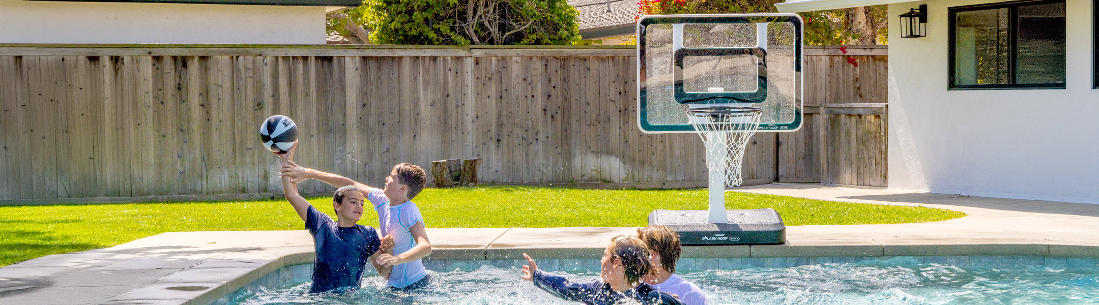 Two children playing with a basketball in a pool, with a basketball hoop in the background.