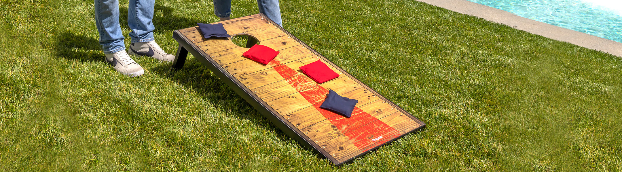 Two people playing cornhole on a grassy area near a pool
