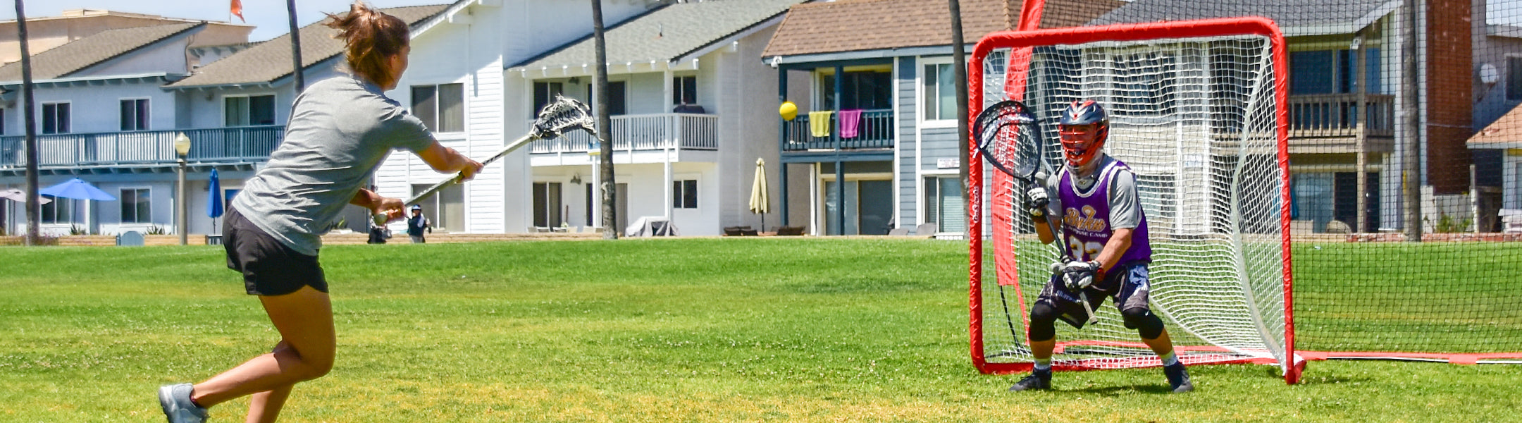Two children playing lacrosse on a grassy field with buildings in the background