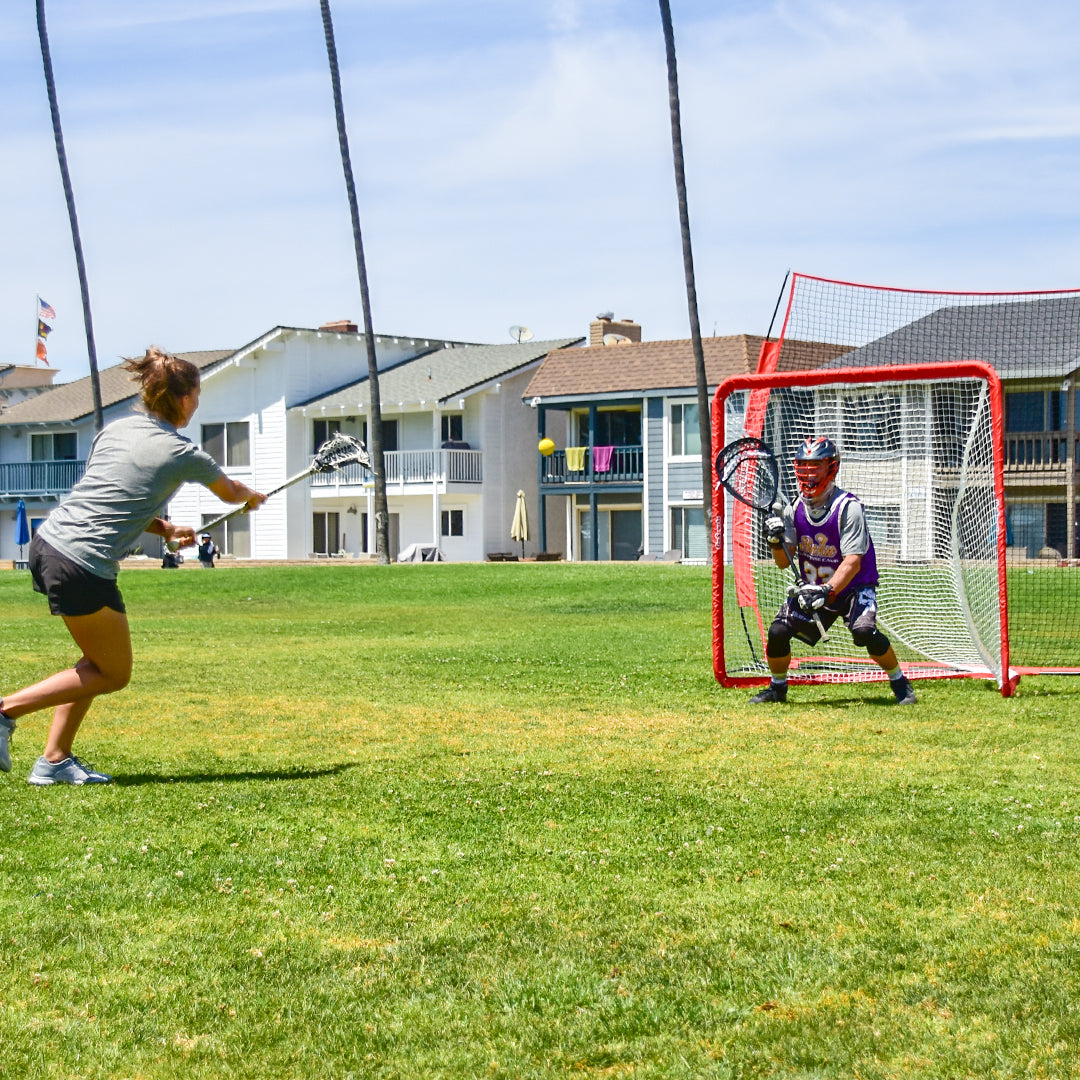 Two people playing lacrosse on a grassy field with houses in the background