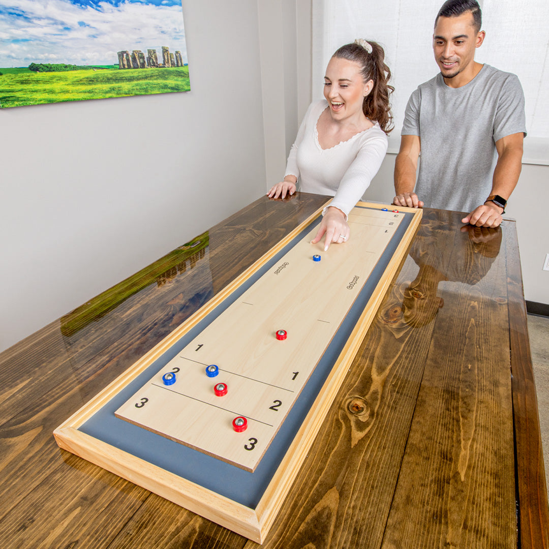 Two people playing a table top shuffle board on a wooden table with a scenic painting in the background.