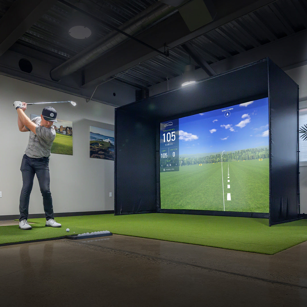 Person practicing golf swings in a simulator room with a screen displaying a golf course.