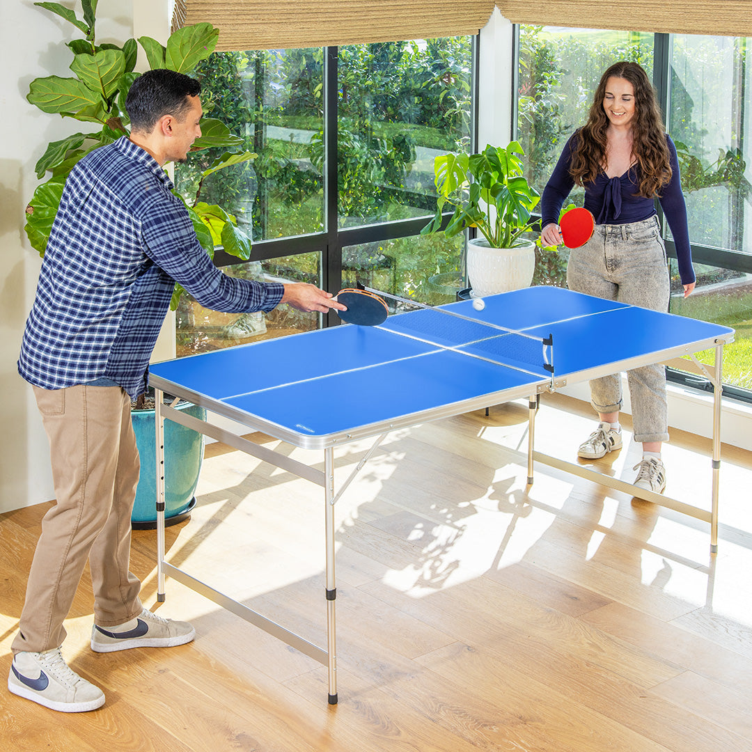 Two people playing table tennis in a room with large windows and plants.