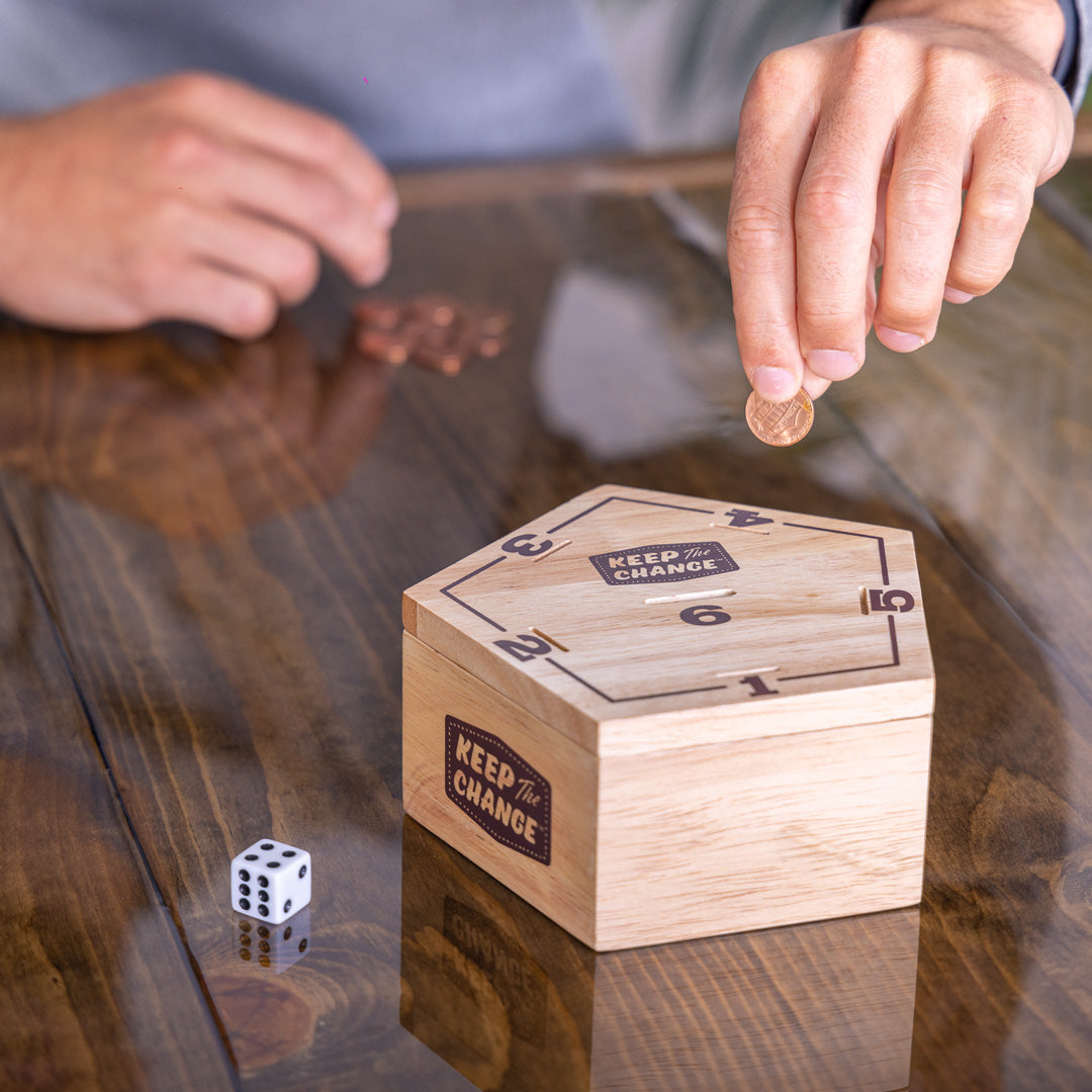 Wooden money box labeled 'Keep the Change' with a hand placing a coin inside on a reflective surface.