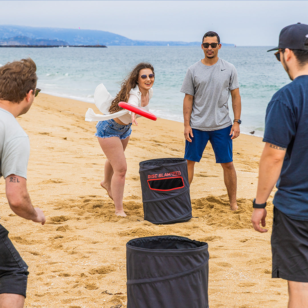 Group of people throwing a frisbee on a beach with two black DISC SLAM cans 
